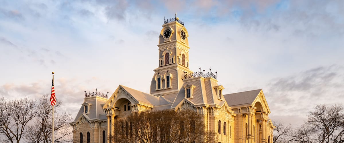 A large, historic building with a prominent clock tower, surrounded by trees and a flagpole.