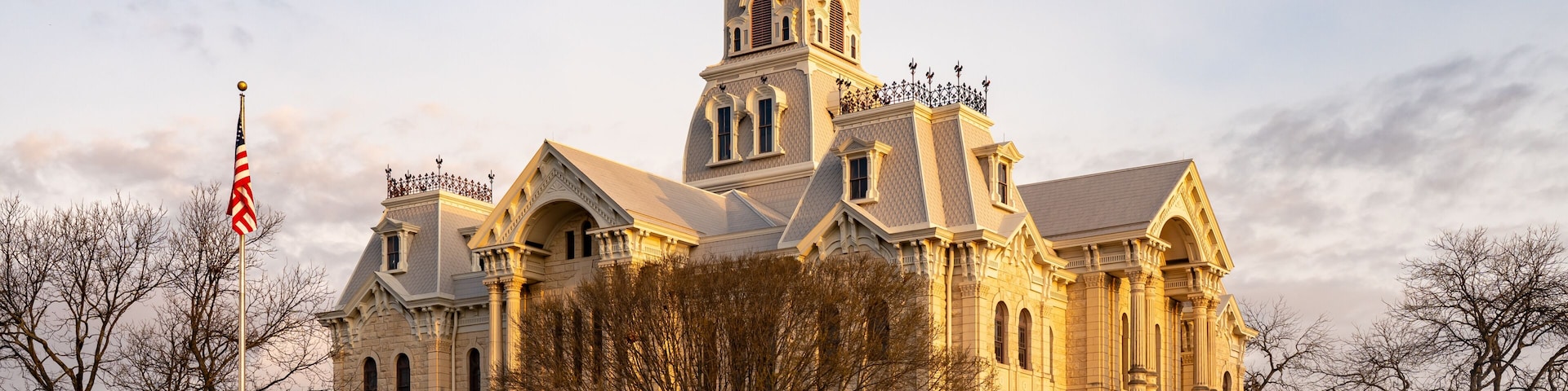 A large, historic building with a prominent clock tower, surrounded by trees and a flagpole.
