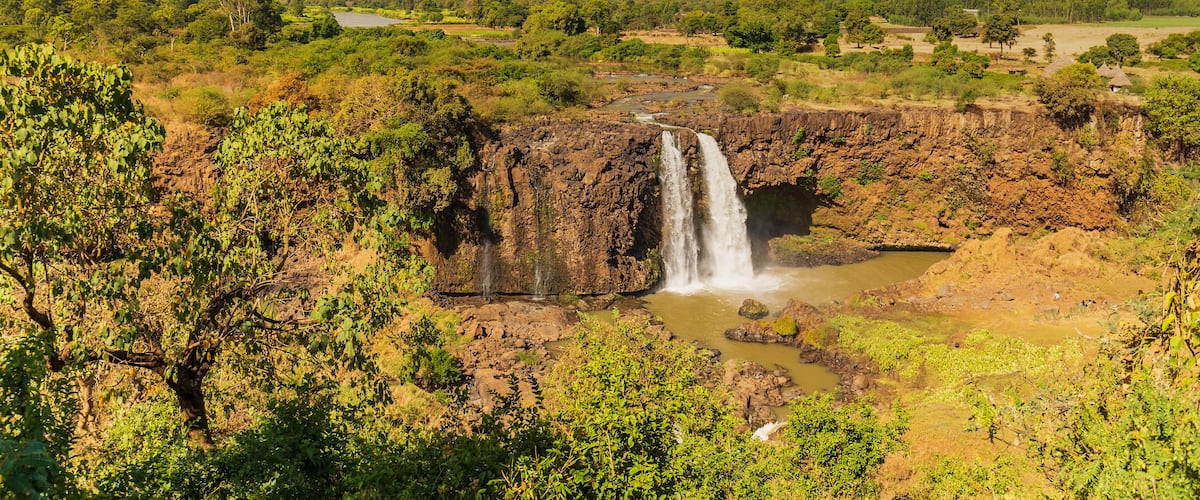 A waterfall cascading into a river surrounded by lush greenery and distant hills.