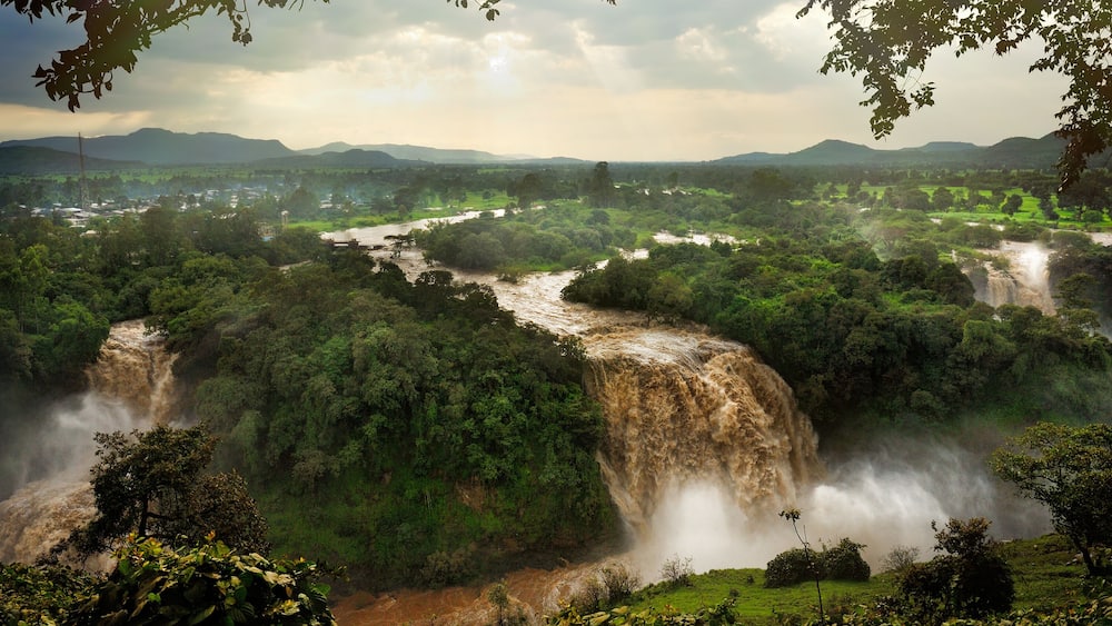 A waterfall cascading through a lush valley surrounded by dense greenery and distant mountains.