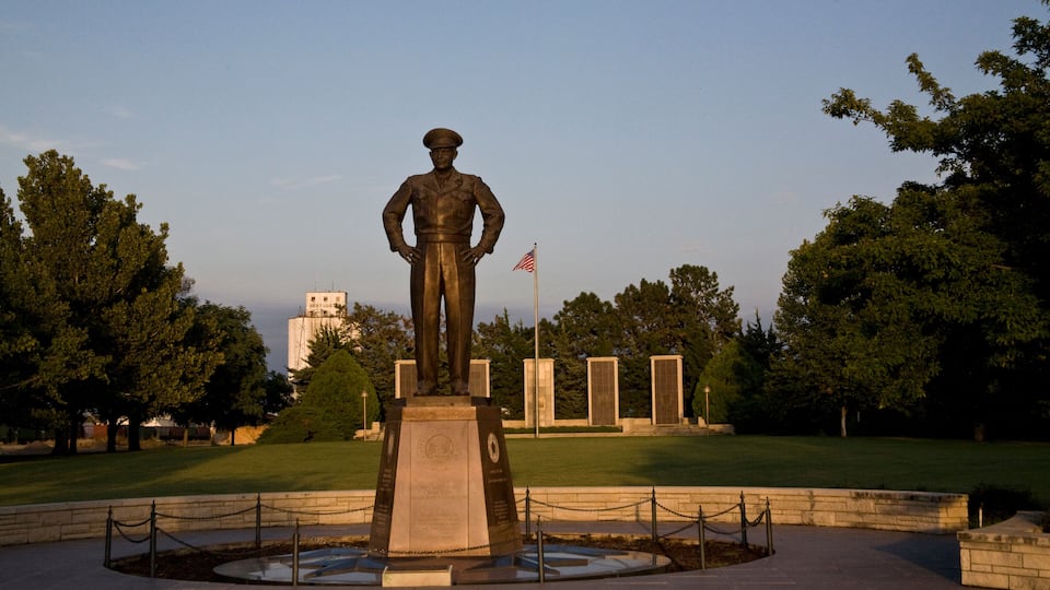 A statue of a soldier with an American flag in a park with trees and a building in the background.