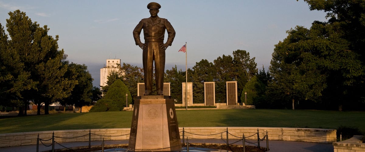A statue of a soldier with an American flag in a park with trees and a building in the background.