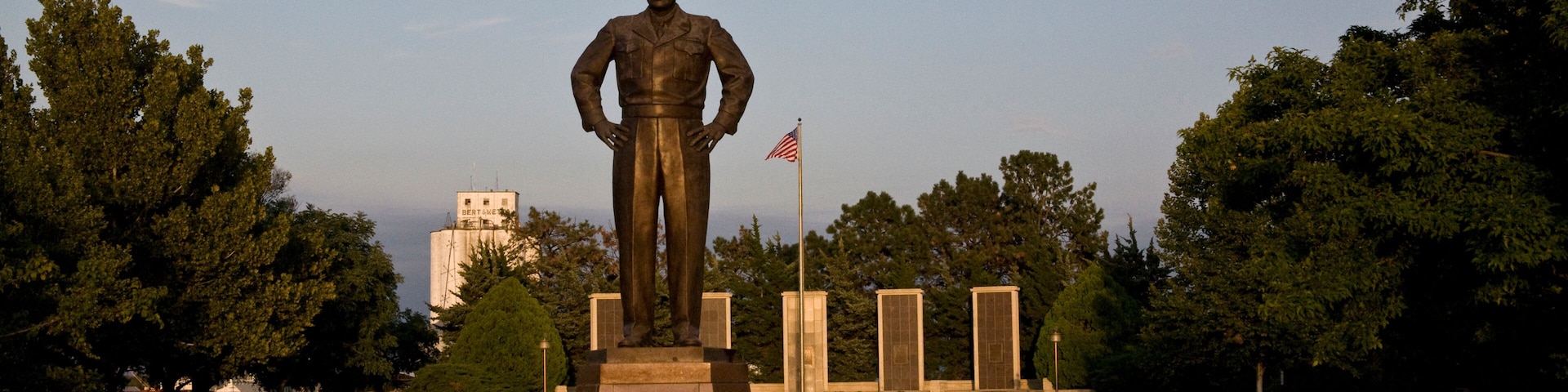 A statue of a soldier with an American flag in a park with trees and a building in the background.
