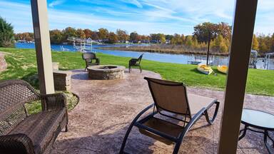 A patio with wicker chairs, a fire pit, and a view of a lake with boats.