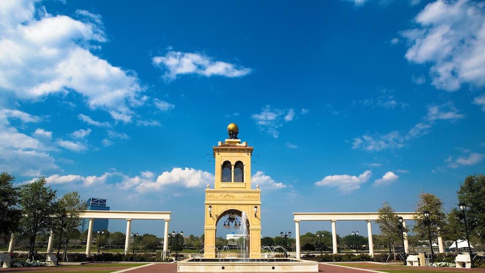 A large yellow tower with a clock and a dome, surrounded by a fountain and a park with trees and a clear blue sky.