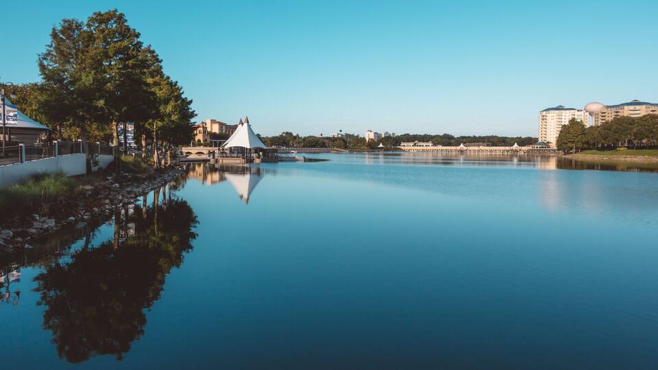 A calm river reflecting a white gazebo, trees, and buildings in the background.