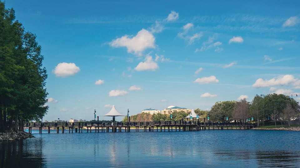 A calm lake with a wooden pier, trees, and a distant building complex.