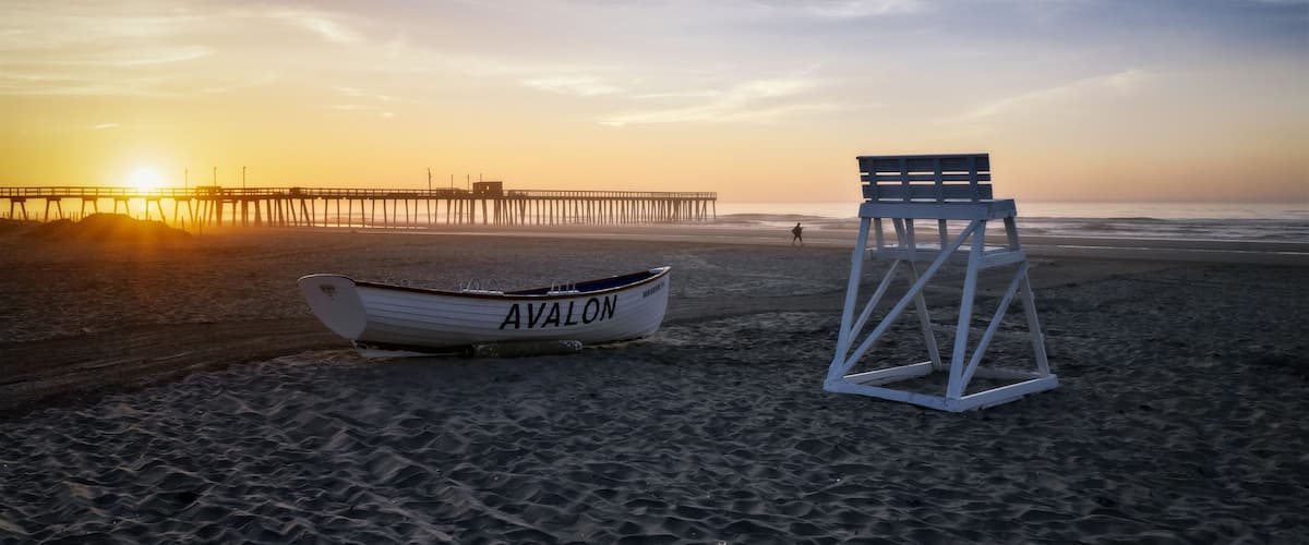 A beach with a lifeguard chair and a boat named 'AVALON' near a pier during sunset.