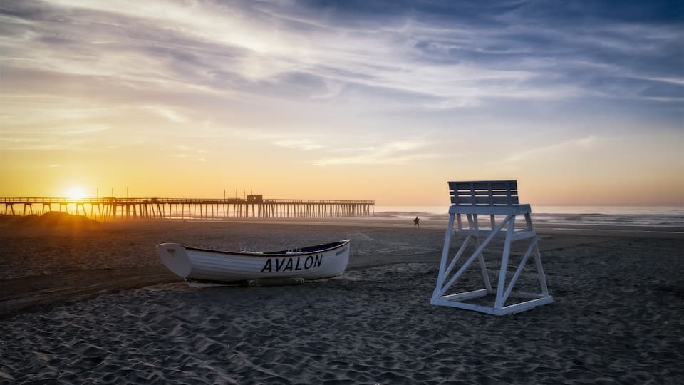 A beach with a lifeguard chair and a boat named 'AVALON' near a pier during sunset.