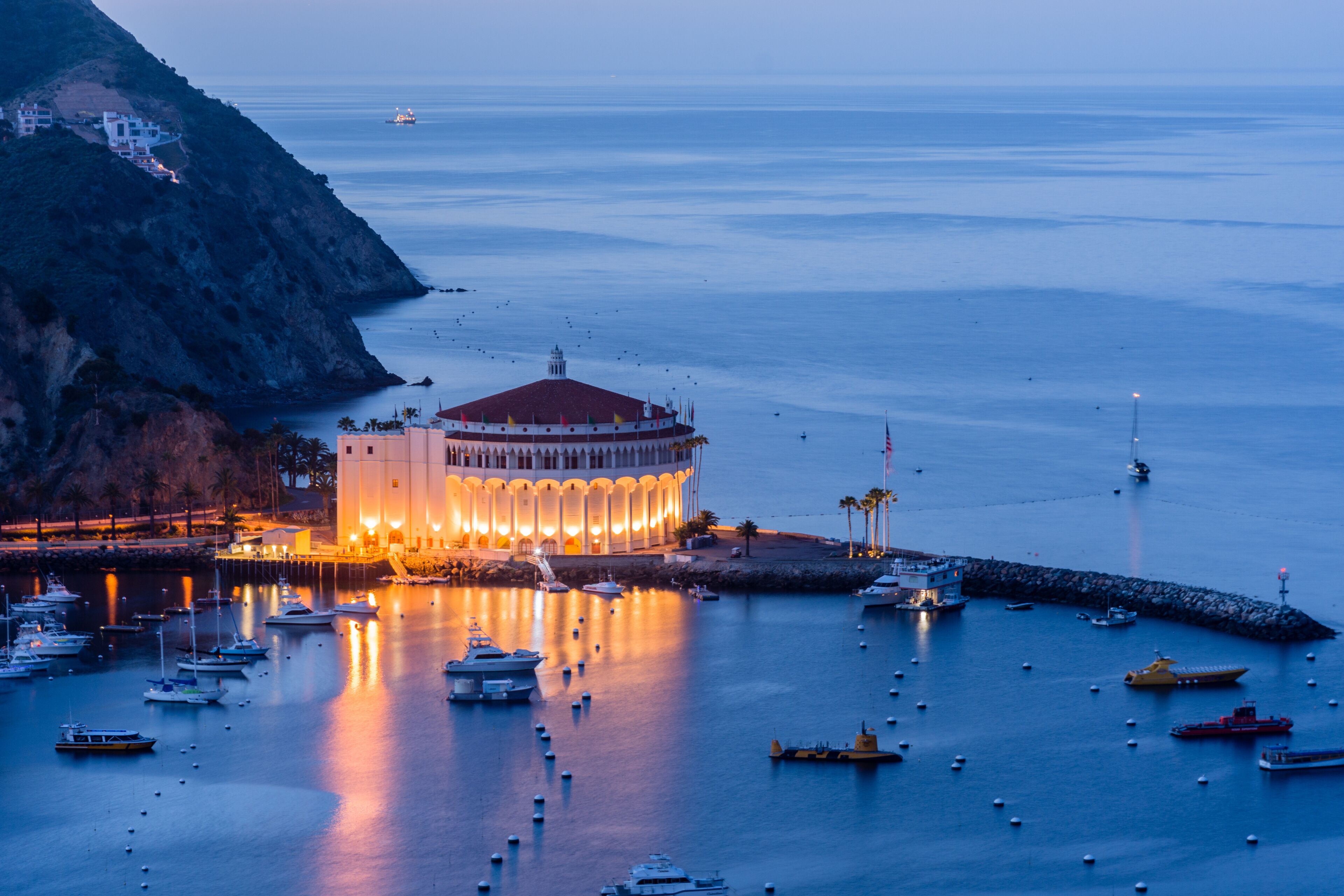 Un pueblo costero con un edificio circular destacado, barcos y un litoral rocoso.