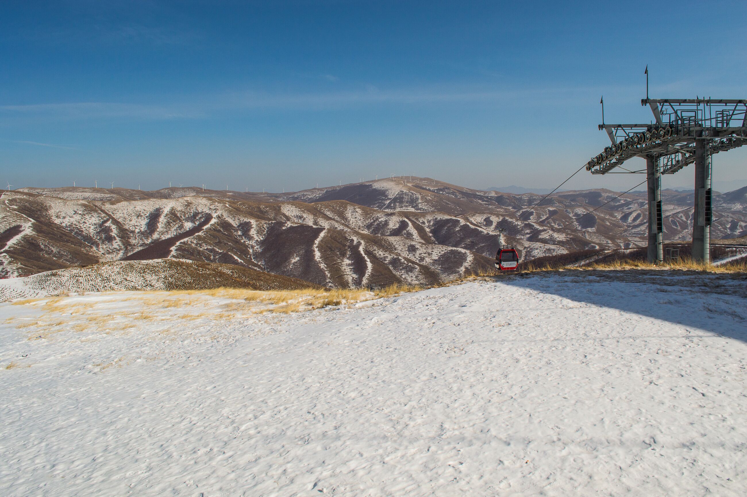 A snowy mountain landscape with a ski lift and wind turbines in the distance.