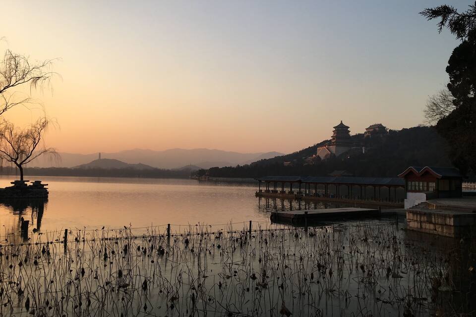 A serene lake at sunset with a traditional pagoda and a pavilion on the right, surrounded by hills and a clear sky.