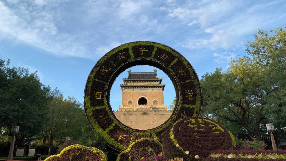 A circular garden with a central view of a traditional gate, surrounded by vibrant flower beds and trees.