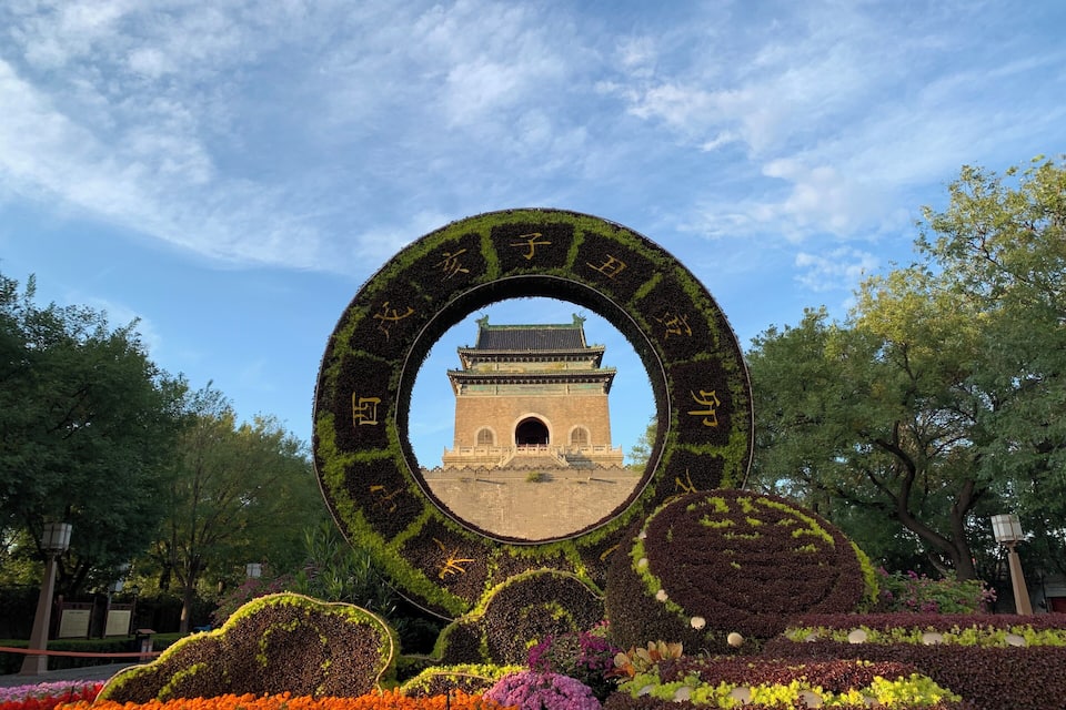 Um jardim circular com vista para um portão tradicional, cercado por canteiros de flores vibrantes e árvores.