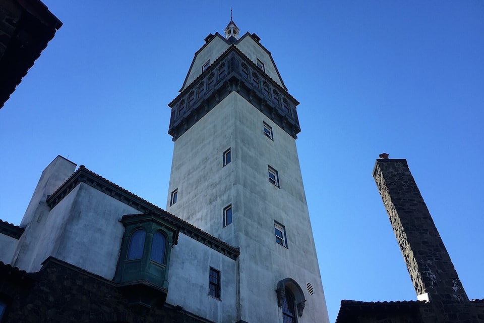 A tall tower with a pointed roof and a clock at the top, surrounded by buildings with stone facades.