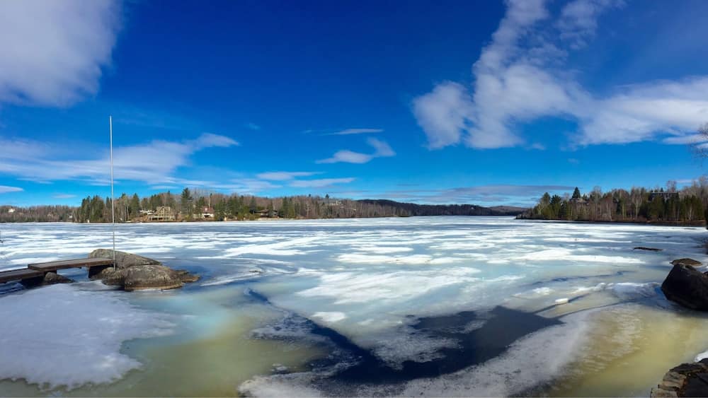 A frozen river with a wooden dock, surrounded by trees and houses under a clear blue sky.