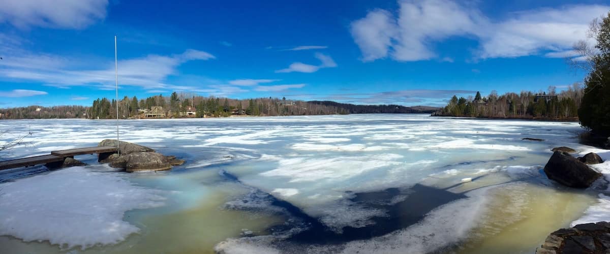 A frozen river with a wooden dock, surrounded by trees and houses under a clear blue sky.