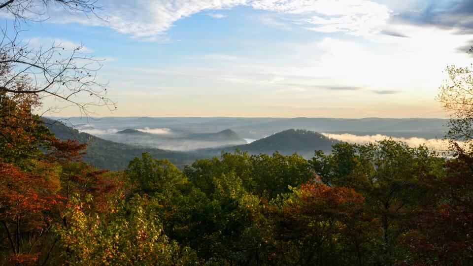 A mountainous landscape with autumn foliage and a view of distant hills under a partly cloudy sky.