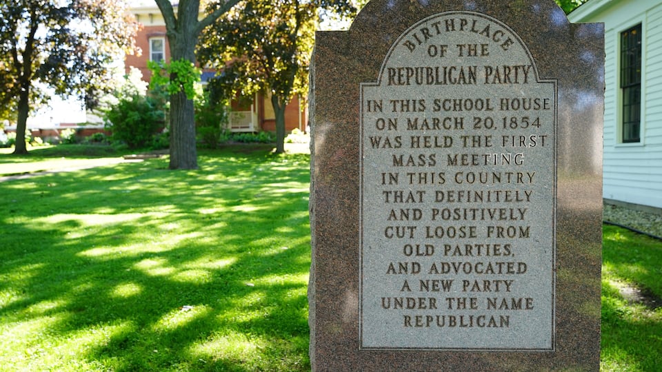 A historical marker in a grassy area with trees and a building in the background.
