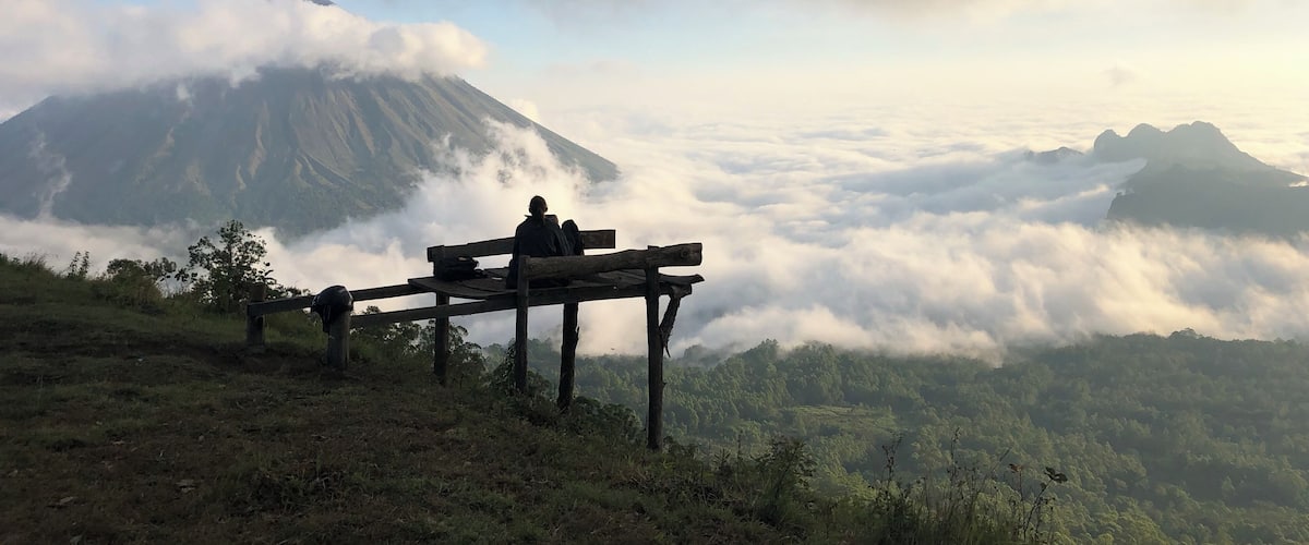 A mountain with a person sitting on a bench, surrounded by clouds and greenery.