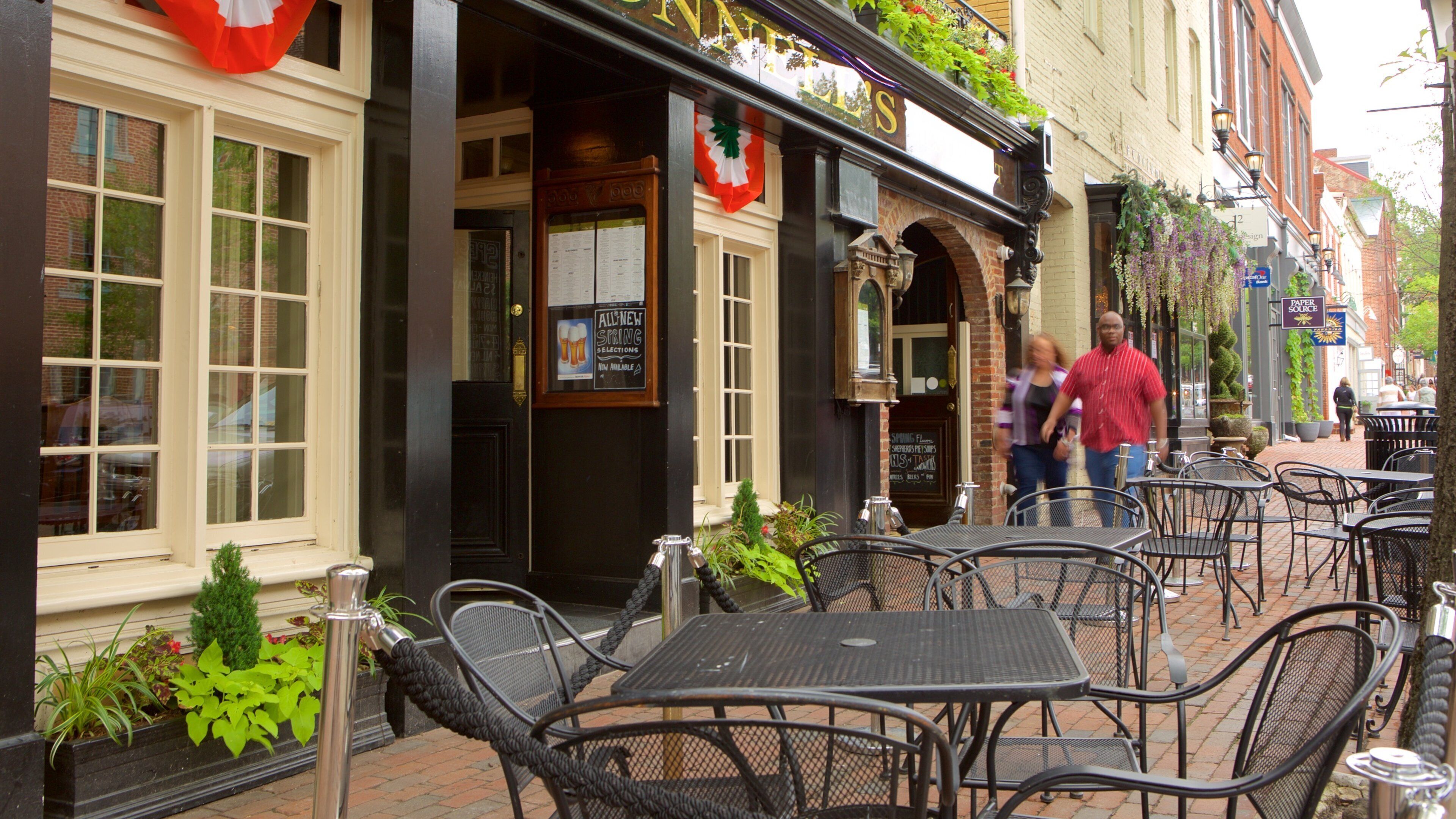Outdoor cafe seating with tables and chairs, a storefront with a sign, and a brick building in the background.