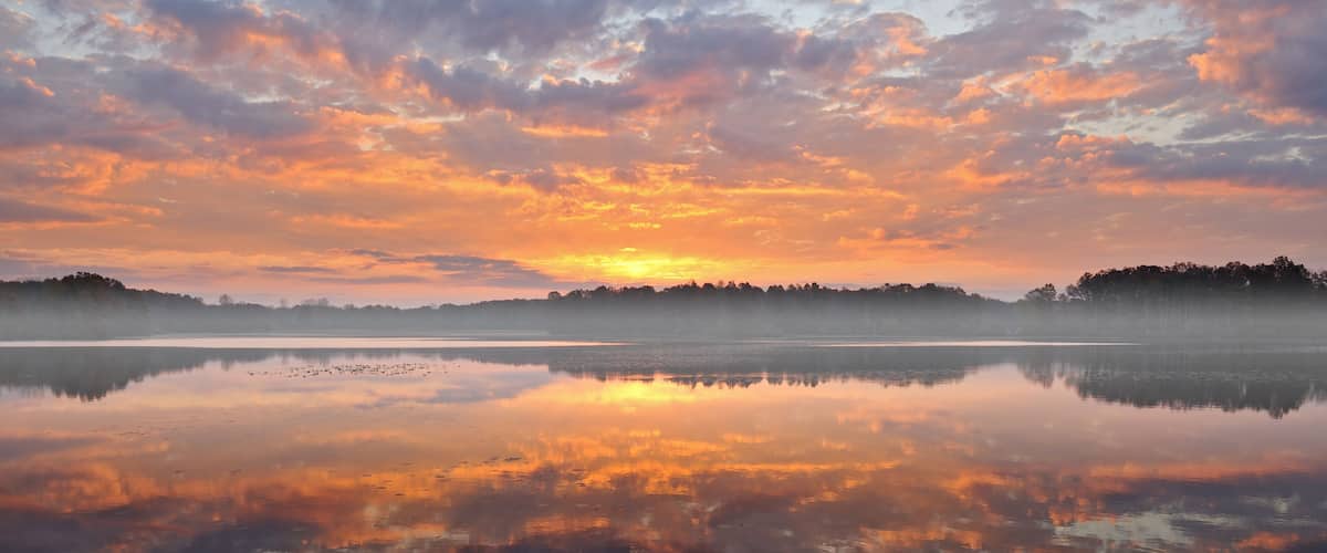 A calm lake reflecting a sunset sky with clouds, surrounded by a forested area.