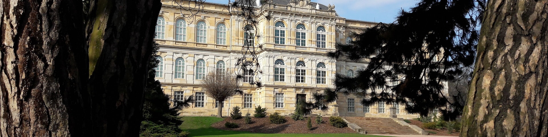 Un edificio grande e histórico con una fachada simétrica, rodeado de un césped cuidado y árboles altos.