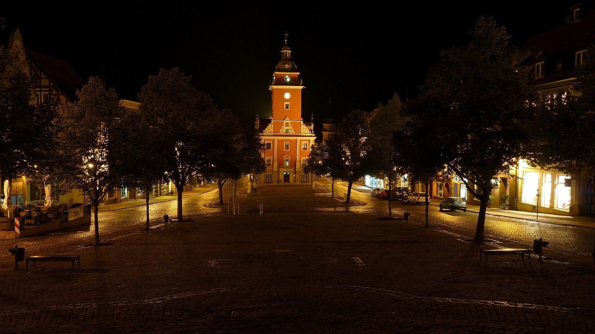 A nighttime view of a square with a prominent clock tower, surrounded by trees and illuminated buildings.