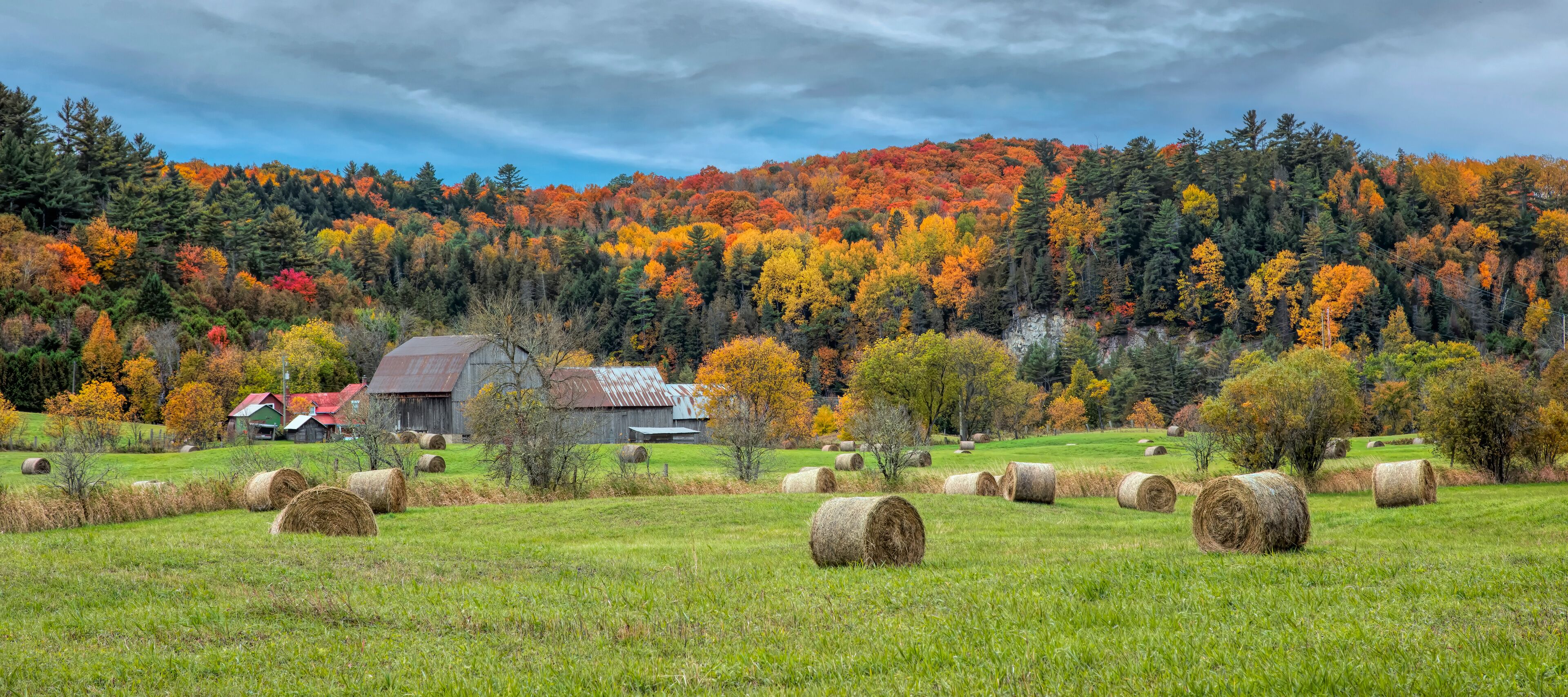 A rural landscape with hay bales, a barn, and autumn-colored trees.