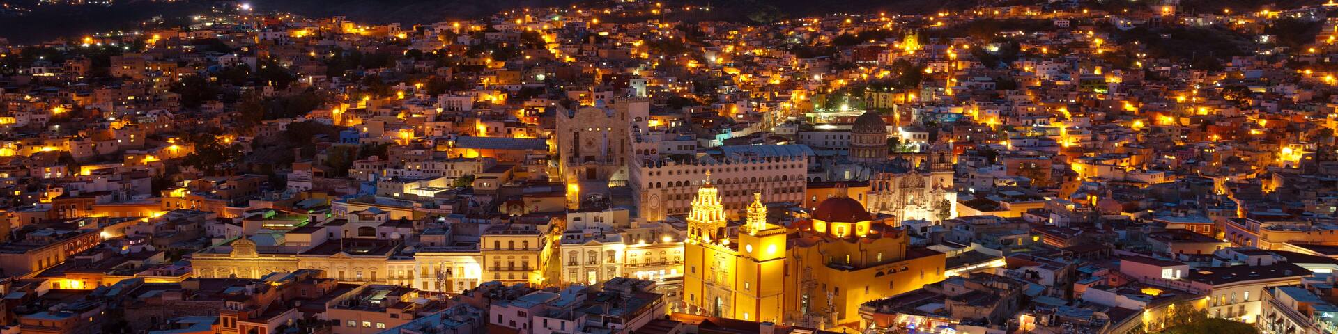 A cityscape at dusk with illuminated buildings and a prominent cathedral with a dome.