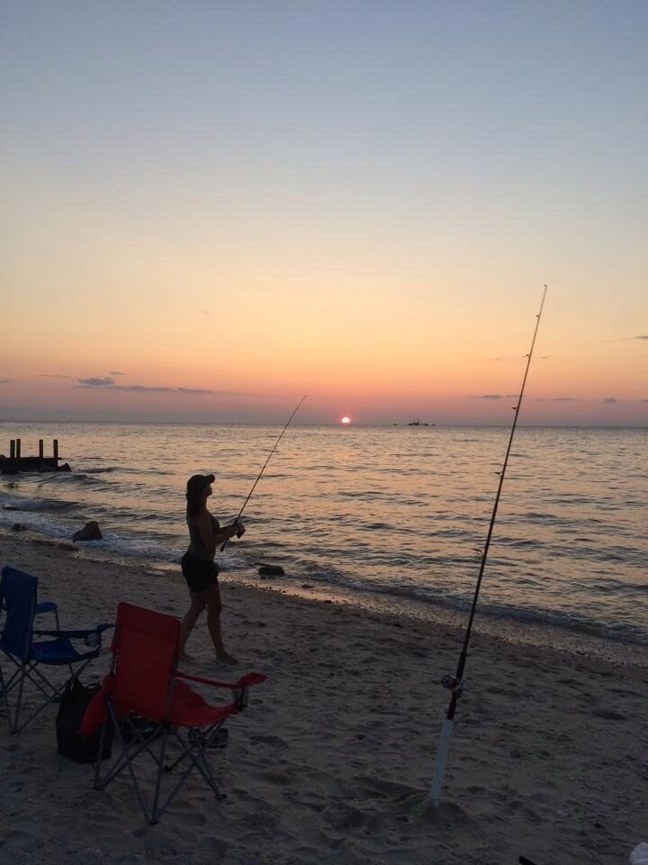 A person fishing on a beach at sunset with chairs and a fishing rod in the foreground.