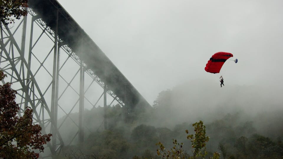 A person skydiving with a red parachute near a bridge and foggy forest.