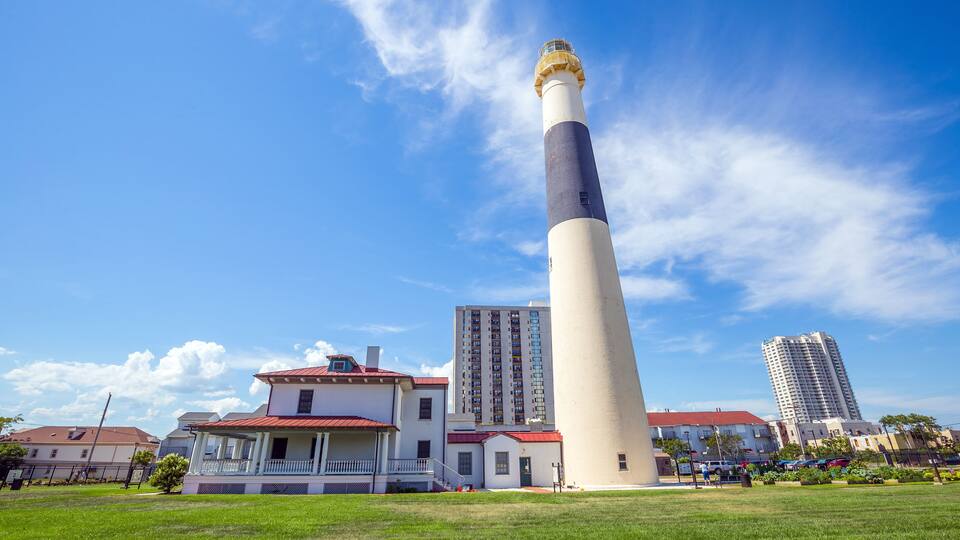 A lighthouse with a white and black tower, a red-roofed building, and a tall residential building in the background.