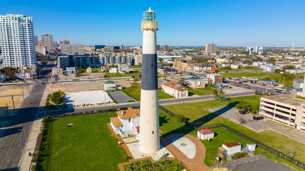 A tall lighthouse with a white and gray tower, surrounded by a well-maintained grassy area, amidst a cityscape with various buildings and a clear blue sky.