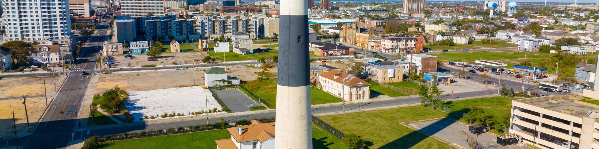 A tall lighthouse with a white and gray tower, surrounded by a well-maintained grassy area, amidst a cityscape with various buildings and a clear blue sky.