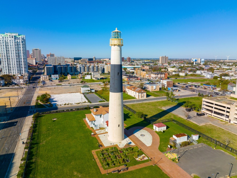 Un faro alto con torre blanca y gris, rodeado de un césped cuidado, en medio de un paisaje urbano con diversos edificios y un cielo azul despejado.