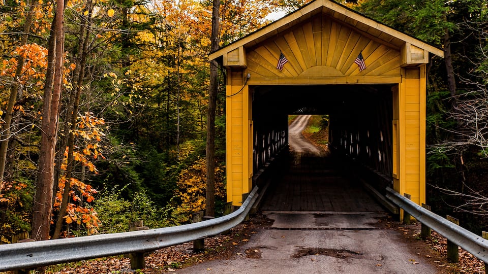 A yellow covered bridge with an American flag, surrounded by autumn foliage.