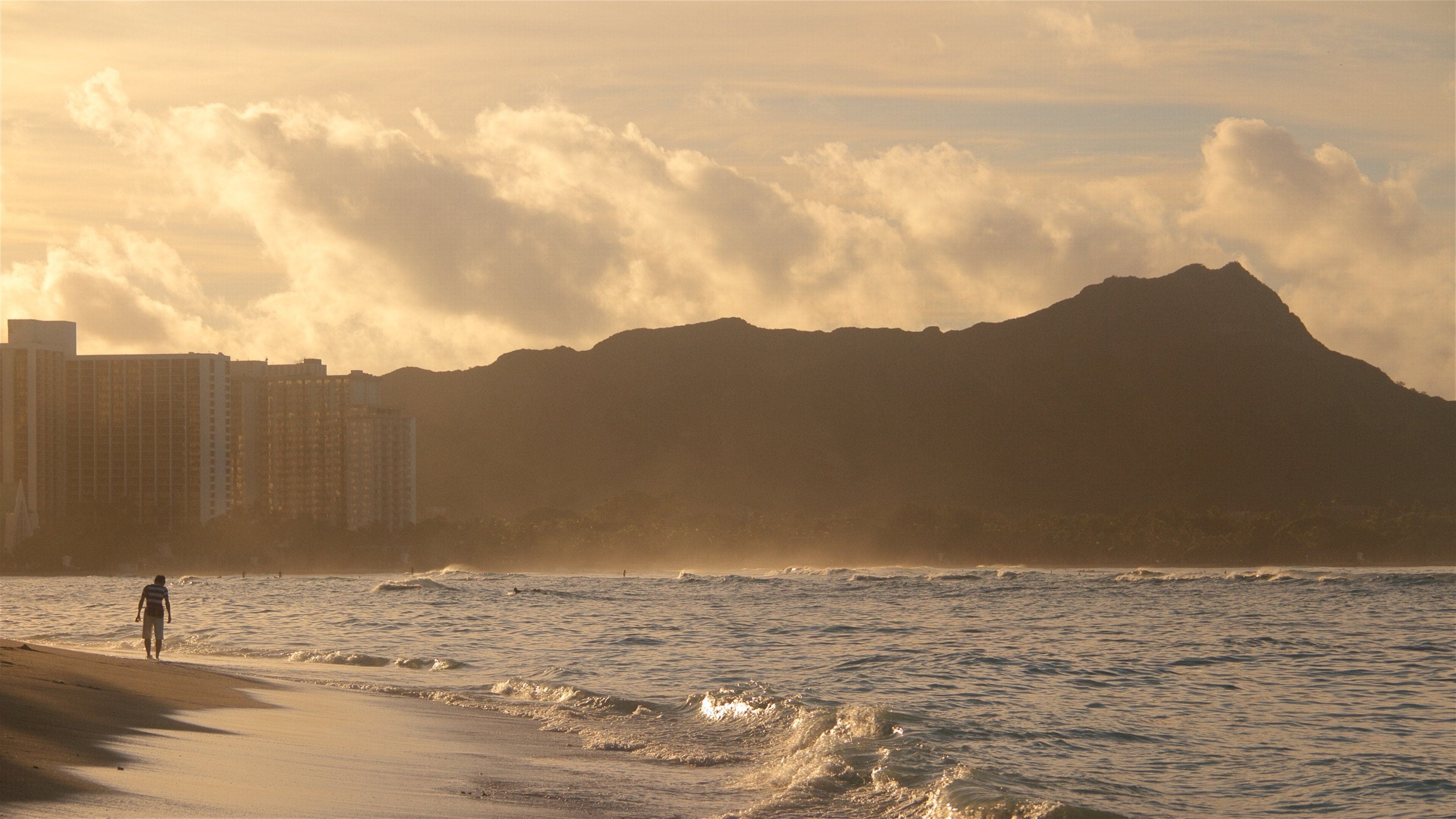 Uma praia com ondas, uma pessoa caminhando e uma montanha ao fundo.