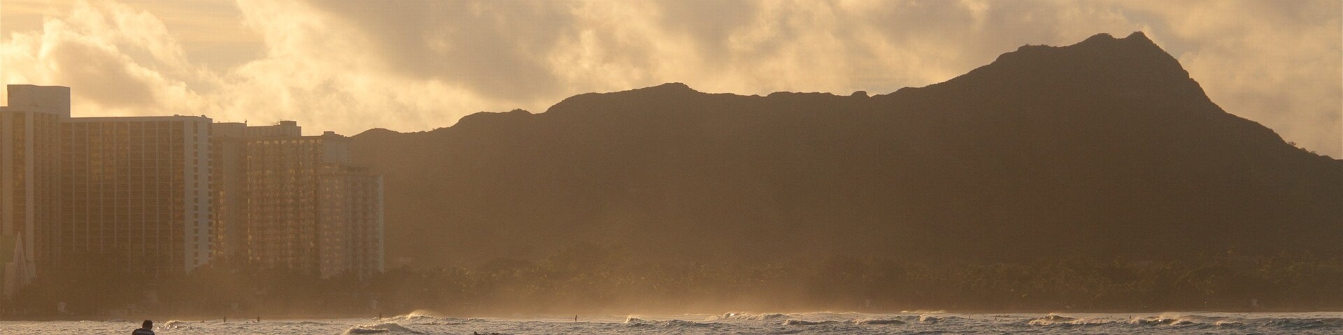 Uma praia com ondas, uma pessoa caminhando e uma montanha ao fundo.