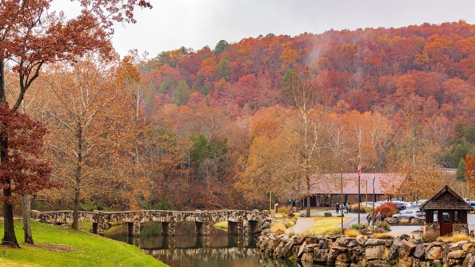 A stone bridge over a calm river, surrounded by autumn-colored trees and a small wooden building.