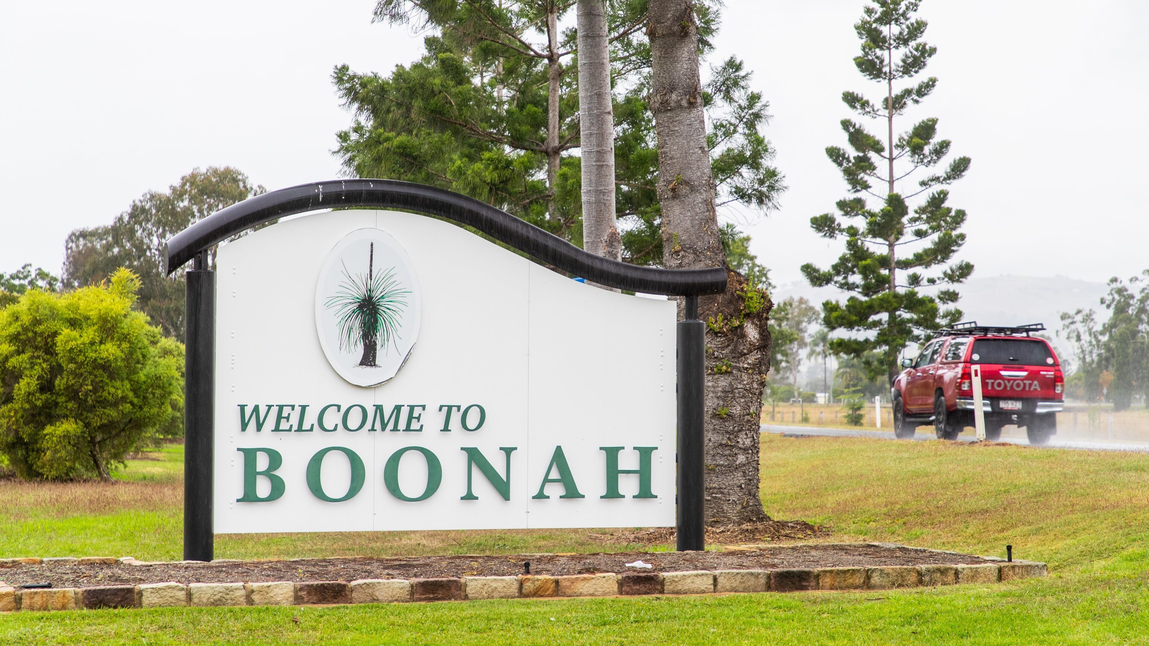 A welcome sign for Boonah with a tree illustration, surrounded by greenery and a red vehicle in the background.