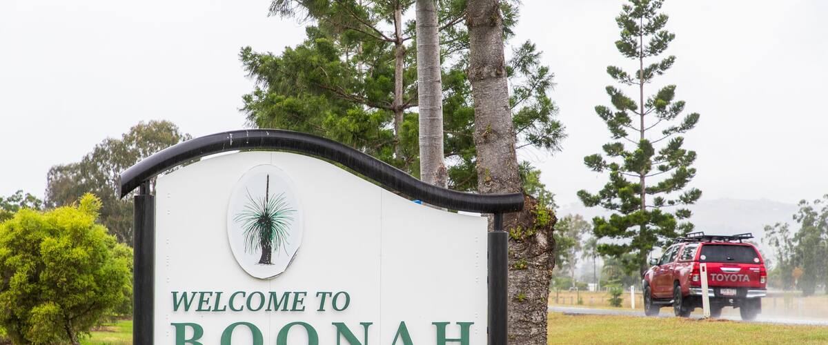 A welcome sign for Boonah with a tree illustration, surrounded by greenery and a red vehicle in the background.