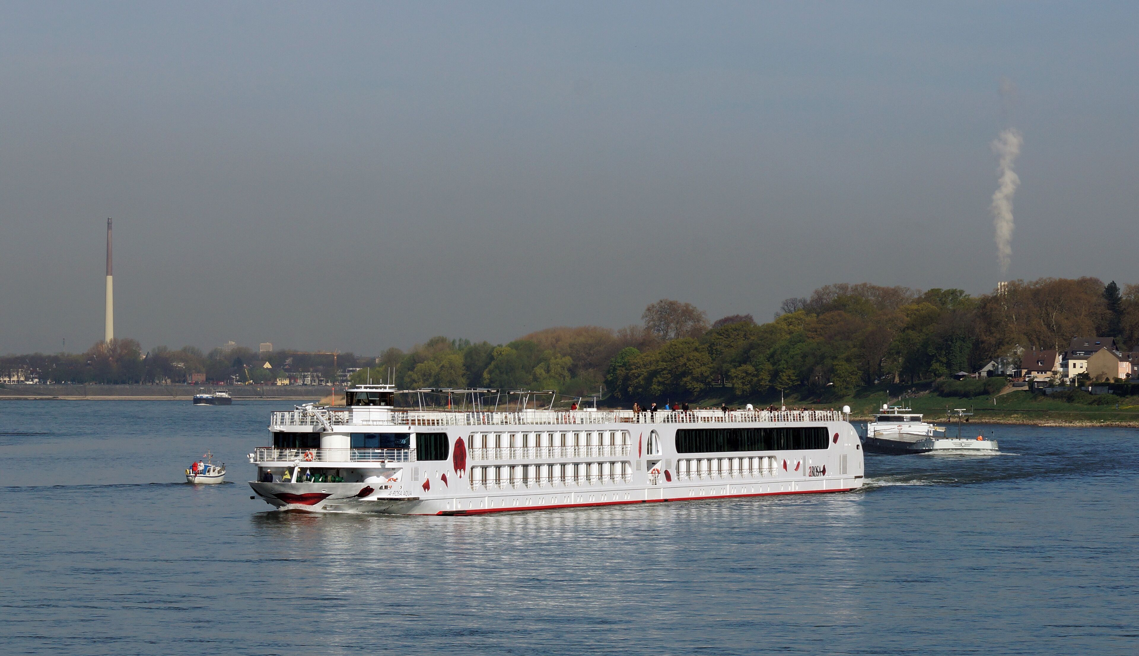 A river cruise ship with passengers, a smaller boat nearby, and industrial chimneys in the background.