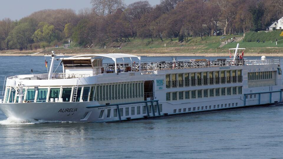 Ein weißes Flusskreuzfahrtschiff namens AURELIA auf einem ruhigen Fluss.