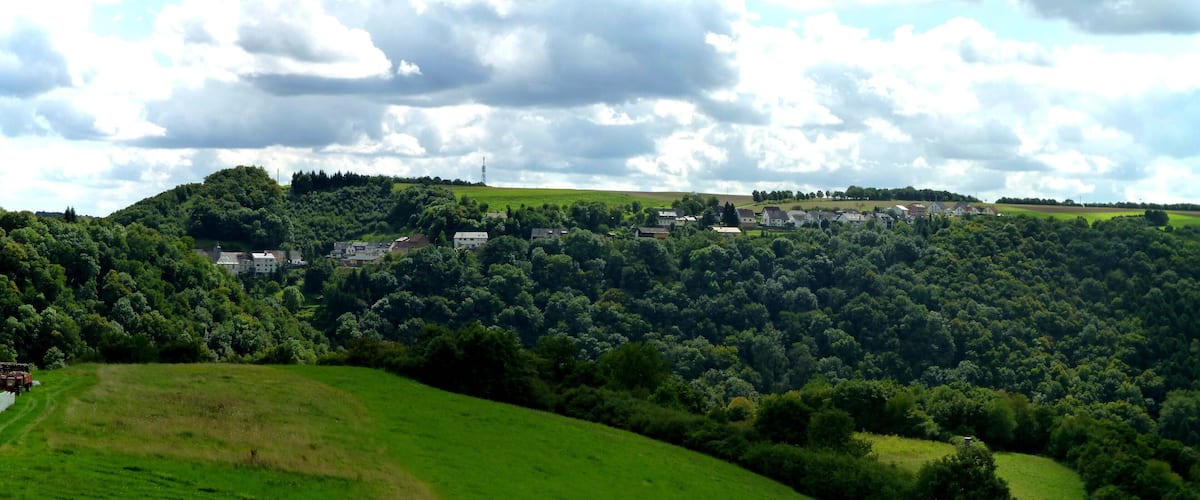 A rural landscape with a village nestled in a valley surrounded by lush green hills and a cloudy sky.