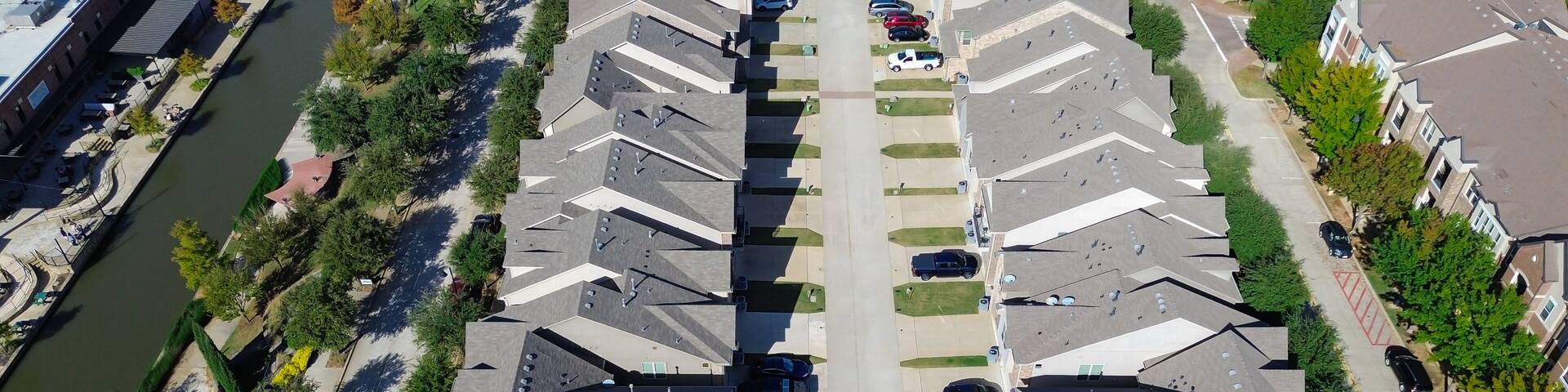 Panorama aerial canal with bridge, waterfall, flat roof restaurant, parking garage next row of suburban homes gabled roofs, integration of residential living, commercial zones, Flower Mound, TX