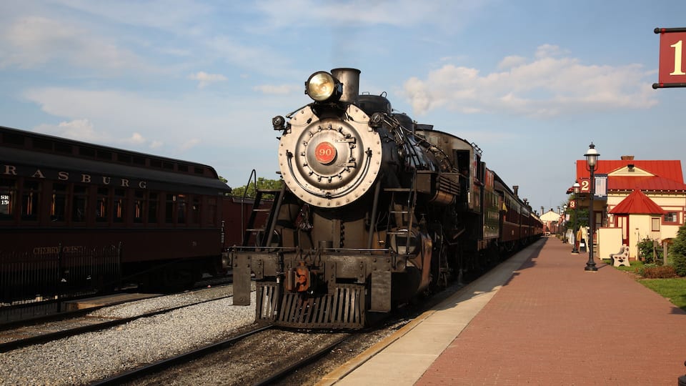 A steam locomotive at a train station with tracks, a platform, and a building with a red roof.