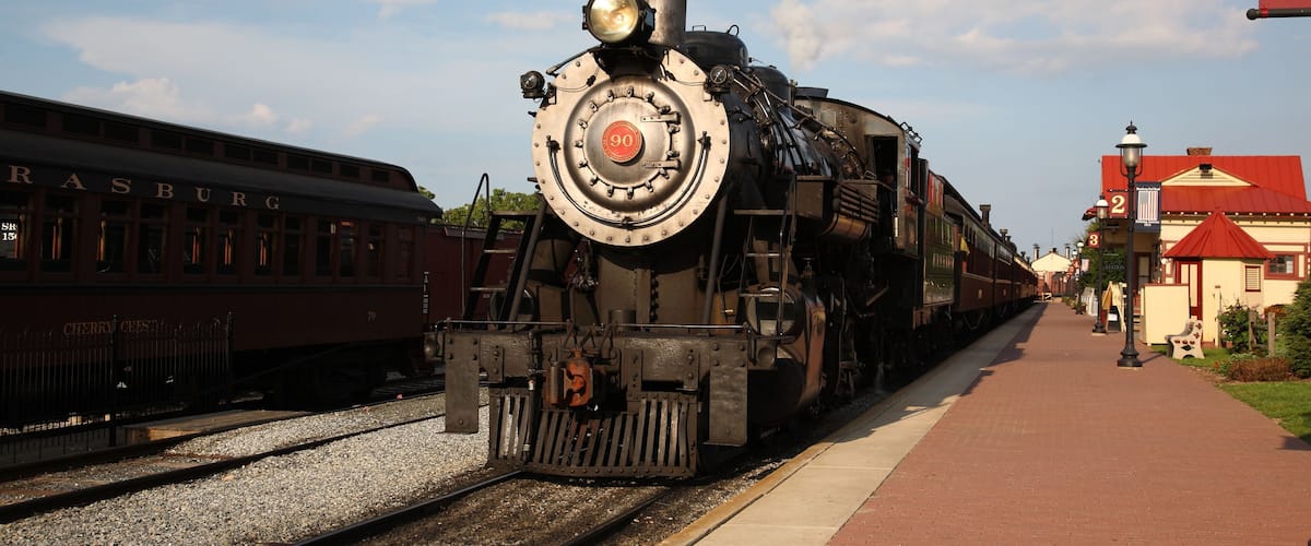 A steam locomotive at a train station with tracks, a platform, and a building with a red roof.
