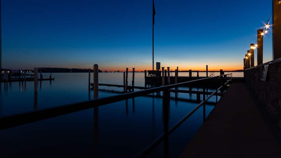 A waterfront at dusk with a dock, a flagpole, and a row of lights.