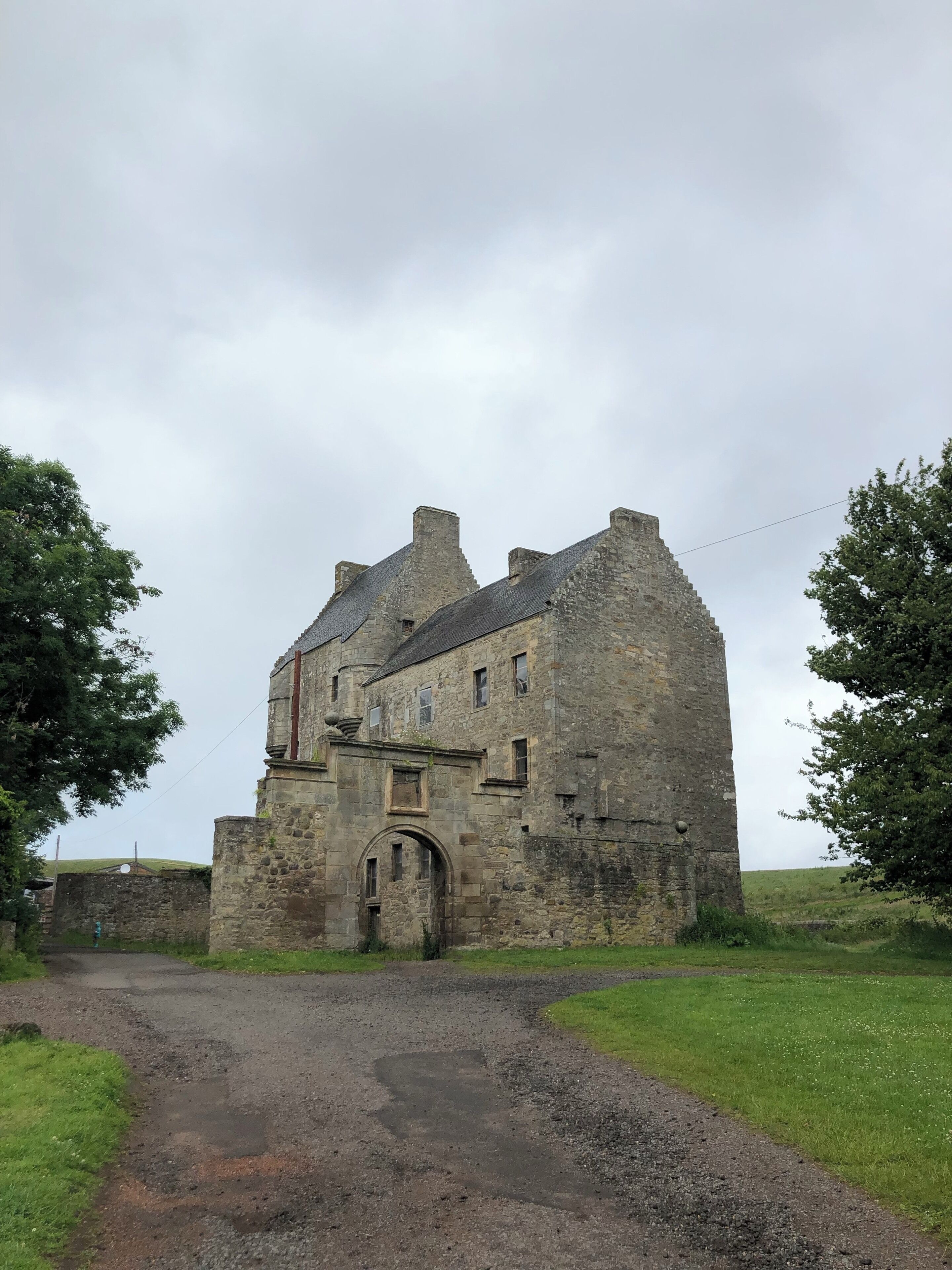 A historic stone building with a large arched entrance, surrounded by grassy fields and trees.